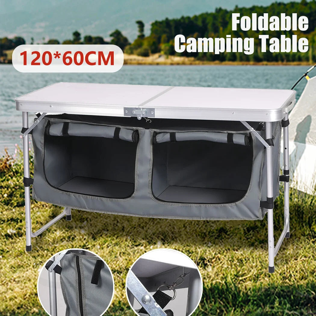 Foldable camping table with storage compartments on a grassy area by a body of water.