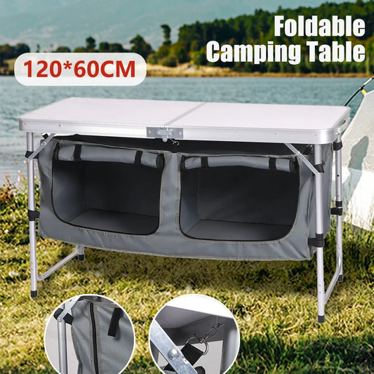 Foldable camping table with storage compartments on a grassy area by a body of water.