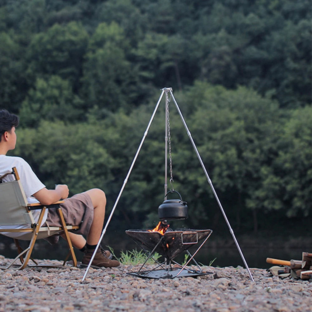 Person sitting by a campfire with a tripod stand holding a pot outdoors.